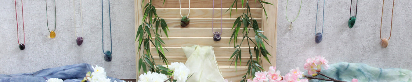 Das Bild zeigt diverse gebohrte Trommelsteine als Ketten an einer weißen Wand. Als Deko liegen bunte Tücher und Kirschblüten auf dem Boden.