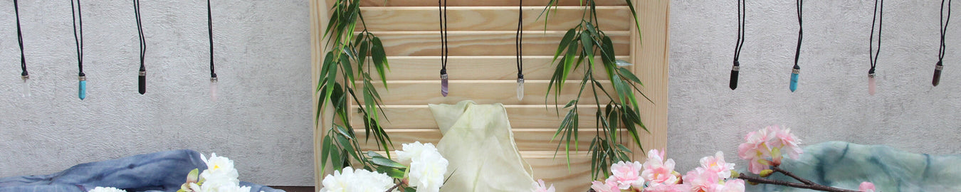 Das Bild zeigt diverse Edelstein Stift-Anhänger mit Kette an einer weißen Wand. Als Deko liegen bunte Tücher und Kirschblüten auf dem Boden.