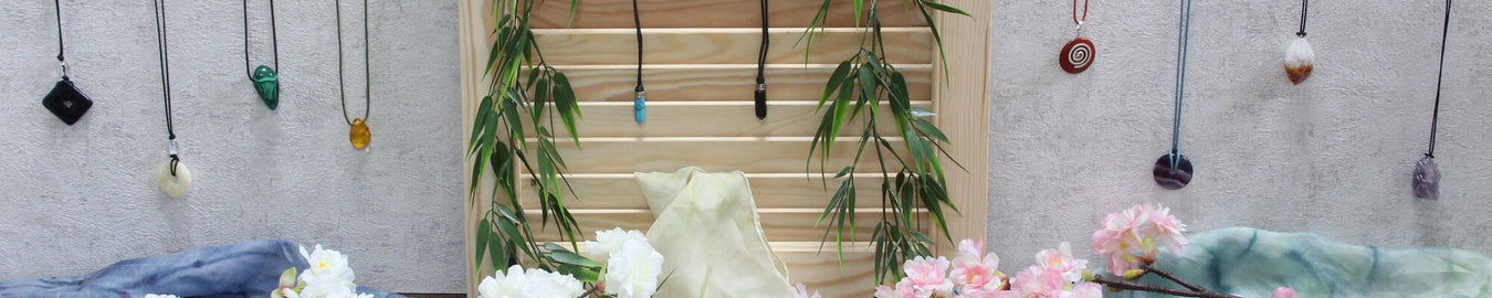 Das Bild zeigt diverse Edelstein-Ketten an einer weißen Wand. Als Deko liegen bunte Tücher und Kirschblüten auf dem Boden.