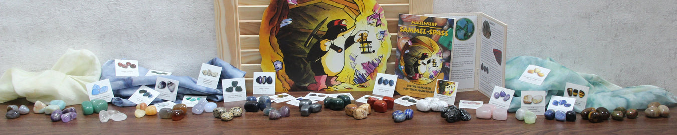 Maulwurf-Sammelspaß Trommelsteine Titelbild. Das Bild zeigt alle 24 Maulwurf Sammelspaßsteine mit den Stickern und dem Sammelheft auf einem hölzernen Untergrund. Als Deko liegen bunte Seidentücher dabei.