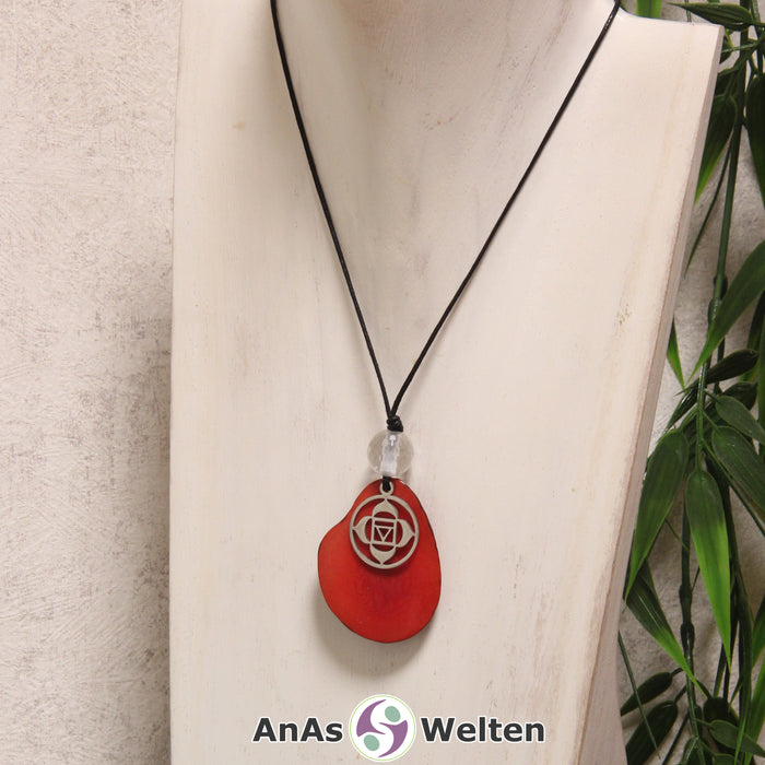 Das Produktbild für die 1. Chakra (Wurzelchakra) Symbol Kette rot zeigt eine rot gefärbte Tagua-Nuss-Scheibe an einem schwarzen Baumwollband, vor der ein silberfarbener Anhänger mit dem Chakrasymbol befestigt ist. Die Nuss-Scheibe hat einen dunklen Rand und eine glänzende Oberfläche. Direkt darüber befindet sich eine Bergkristallkugel.