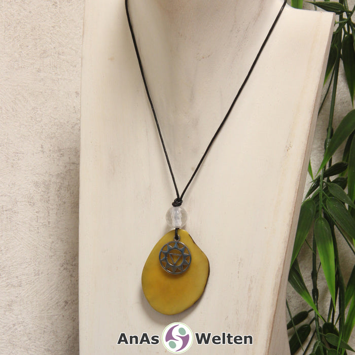 Das Produktbild für die 3. Chakra (Solarplexuschakra) Symbol Kette gelb zeigt eine gelb gefärbte Tagua-Nuss-Scheibe an einem schwarzen Baumwollband, vor der ein silberfarbener Anhänger mit dem Chakrasymbol befestigt ist. Die Nuss-Scheibe hat einen dunklen Rand und eine glänzende Oberfläche. Direkt darüber befindet sich eine Bergkristallkugel.