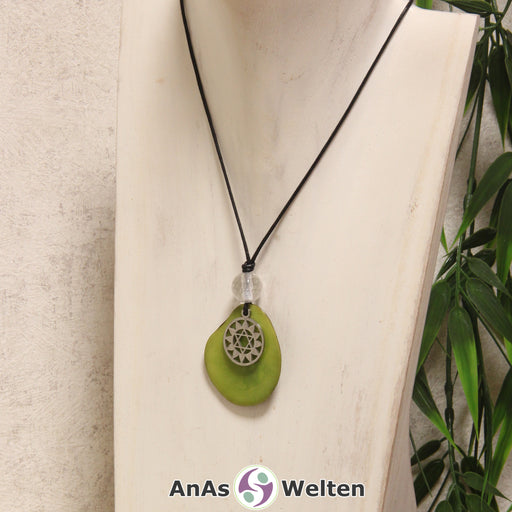 Das Produktbild für die 4. Chakra (Halschakra) Symbol Kette grün zeigt eine grün gefärbte Tagua-Nuss-Scheibe an einem schwarzen Baumwollband, vor der ein silberfarbener Anhänger mit dem Chakrasymbol befestigt ist. Die Nuss-Scheibe hat einen dunklen Rand und eine glänzende Oberfläche. Direkt darüber befindet sich eine Bergkristallkugel.