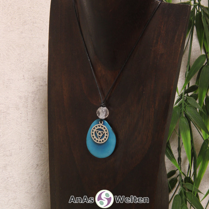 Eine türkise Tagua-Nuss Chakra Kette mit silberfarbenem 5. Chakra Symbol und Bergkristallkugel an einer dunklen Büste.
