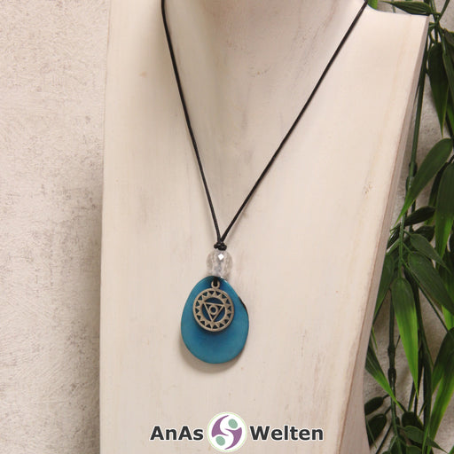 Das Produktbild für die 5. Chakra (Halschakra) Symbol Kette türkis zeigt eine türkis gefärbte Tagua-Nuss-Scheibe an einem schwarzen Baumwollband, vor der ein silberfarbener Anhänger mit dem Chakrasymbol befestigt ist. Die Nuss-Scheibe hat einen dunklen Rand und eine glänzende Oberfläche. Direkt darüber befindet sich eine Bergkristallkugel.