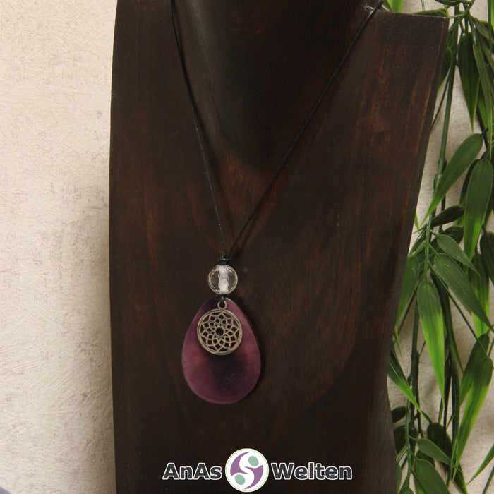 Eine lila Tagua-Nuss Chakra Kette mit silberfarbenem 7. Chakra Symbol und Bergkristallkugel an einer dunklen Büste.