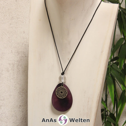 Das Produktbild für die 7. Chakra (Kronenchakra) Symbol Kette violett zeigt eine lila gefärbte Tagua-Nuss-Scheibe an einem schwarzen Baumwollband, vor der ein silberfarbener Anhänger mit dem Chakrasymbol befestigt ist. Die Nuss-Scheibe hat einen dunklen Rand und eine glänzende Oberfläche. Direkt darüber befindet sich eine Bergkristallkugel.