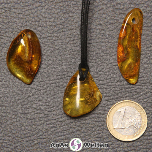 Drei gold-braune Bernstein gebohrte Trommelsteine nebeneinander. Einer befindet sich an einem schwarzen gewachsten Baumwollband.