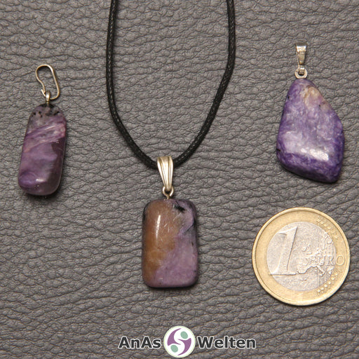 Drei lila Charoit Steine mit Öse nebeneinander. Einer befindet sich an einem schwarzen gewachsten Baumwollband.