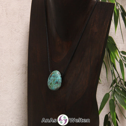 Ein gebohrter Chrysokoll Trommelstein an einem Baumwollband. Der Chrysokoll hat eine türkis-blaue Farbe mit vielen schwarzen, braunen und rotbraunen Einschlüssen, die ihm sein besonderes Muster verleihen.