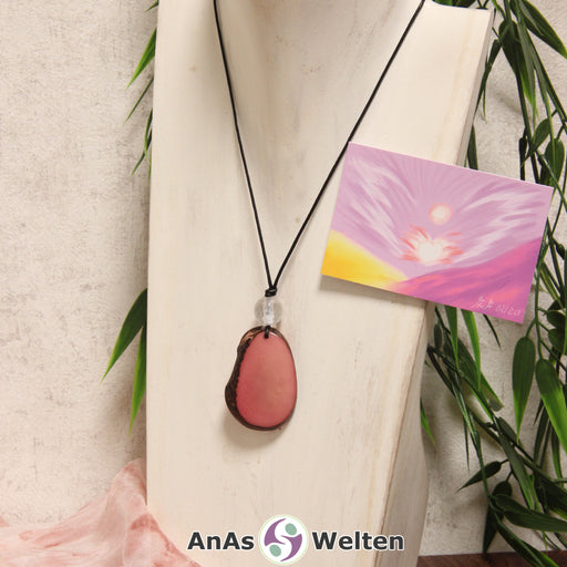 Das Produktbild für die Engel Kette Erzengel Chamuel mit Karte zeigt eine in einem zarten Rosa gefärbte Tagua-Nuss-Scheibe an einem schwarzen Baumwollband. Die Nuss-Scheibe hat einen dunklen Rand und eine glänzende Oberfläche. Direkt darüber befindet sich eine facettierte Bergkristallkugel. Rechts neben der Kette sieht man außerdem die Erzengel Chamuel Karte, die den Engel als abstraktes Bild zeigt.
