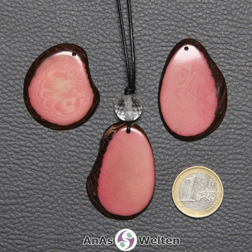 Drei Tagua-Nuss-Scheiben in Rosa. Die mittlere ist mit einer Bergkristallkugel an ein schwarzes Baumwollband gebunden.