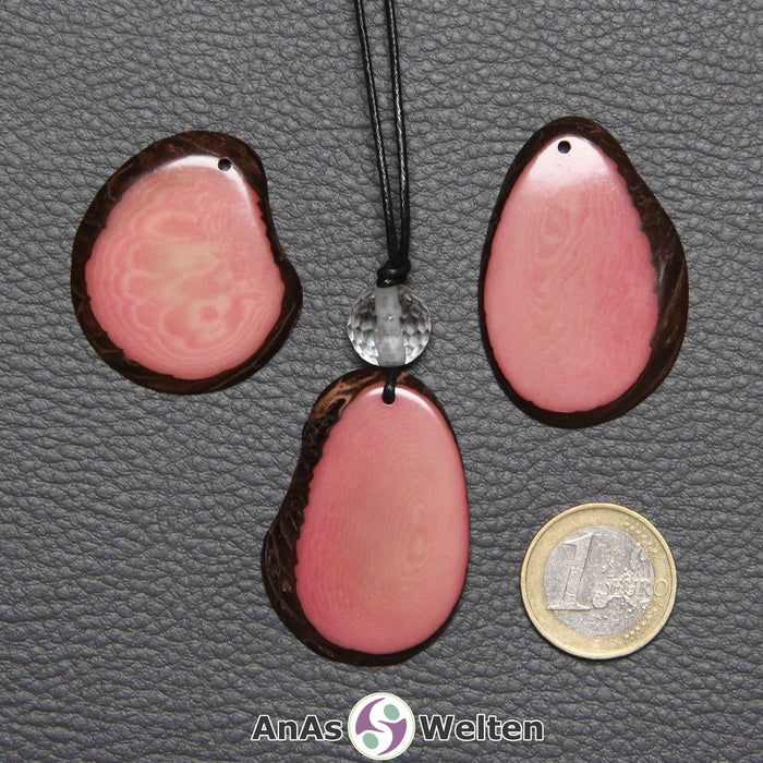 Drei Tagua-Nuss-Scheiben in Rosa. Die mittlere ist mit einer Bergkristallkugel an ein schwarzes Baumwollband gebunden.