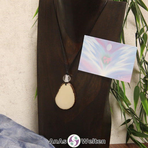 Das Produktbild für die Engel Kette Erzengel Gabriel mit Karte zeigt eine Tagua-Nuss-Scheibe in ihrem natürlichen Cremeweiß an einem schwarzen Baumwollband. Die Nuss-Scheibe hat einen dunklen Rand und eine glänzende Oberfläche. Direkt darüber befindet sich eine facettierte Bergkristallkugel. Rechts neben der Kette sieht man außerdem die Erzengel Gabriel Karte, die den Engel als abstraktes Bild zeigt.