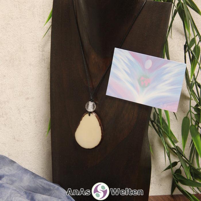 Das Produktbild für die Engel Kette Erzengel Gabriel mit Karte zeigt eine Tagua-Nuss-Scheibe in ihrem natürlichen Cremeweiß an einem schwarzen Baumwollband. Die Nuss-Scheibe hat einen dunklen Rand und eine glänzende Oberfläche. Direkt darüber befindet sich eine facettierte Bergkristallkugel. Rechts neben der Kette sieht man außerdem die Erzengel Gabriel Karte, die den Engel als abstraktes Bild zeigt.