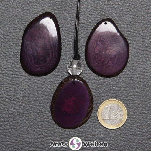 Drei Tagua-Nuss-Scheiben in Lila. Die mittlere ist mit einer Bergkristallkugel an ein schwarzes Baumwollband gebunden.