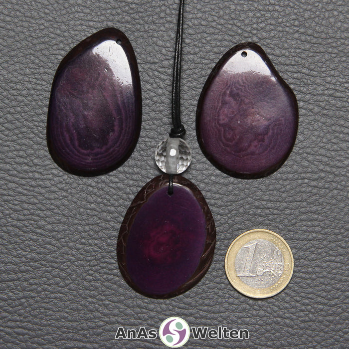 Drei Tagua-Nuss-Scheiben in Lila. Die mittlere ist mit einer Bergkristallkugel an ein schwarzes Baumwollband gebunden.