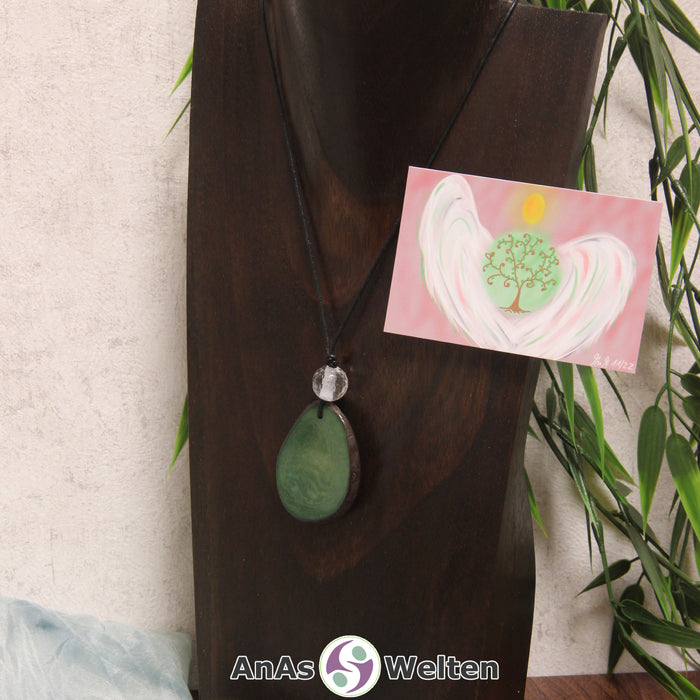 Ein grüner Tagua-Nuss Engel Anhänger ist als Kette mit einer Bergkristallkugel um eine dunkle Büste gehängt.