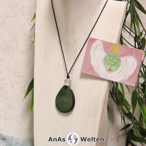 Das Produktbild für die Engel Kette Erzengel Metatron mit Karte zeigt eine in einem dunklen Grün gefärbte Tagua-Nuss-Scheibe an einem schwarzen Baumwollband. Die Nuss-Scheibe hat einen dunklen Rand und eine glänzende Oberfläche. Direkt darüber befindet sich eine facettierte Bergkristallkugel. Rechts neben der Kette sieht man außerdem die Erzengel Metatron Karte, die den Engel als abstraktes Bild zeigt.