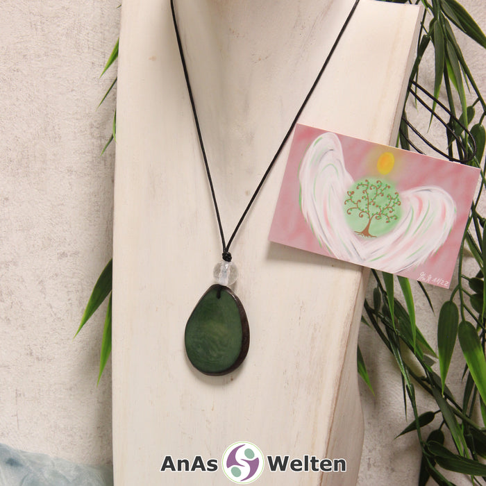 Das Produktbild für die Engel Kette Erzengel Metatron mit Karte zeigt eine in einem dunklen Grün gefärbte Tagua-Nuss-Scheibe an einem schwarzen Baumwollband. Die Nuss-Scheibe hat einen dunklen Rand und eine glänzende Oberfläche. Direkt darüber befindet sich eine facettierte Bergkristallkugel. Rechts neben der Kette sieht man außerdem die Erzengel Metatron Karte, die den Engel als abstraktes Bild zeigt.