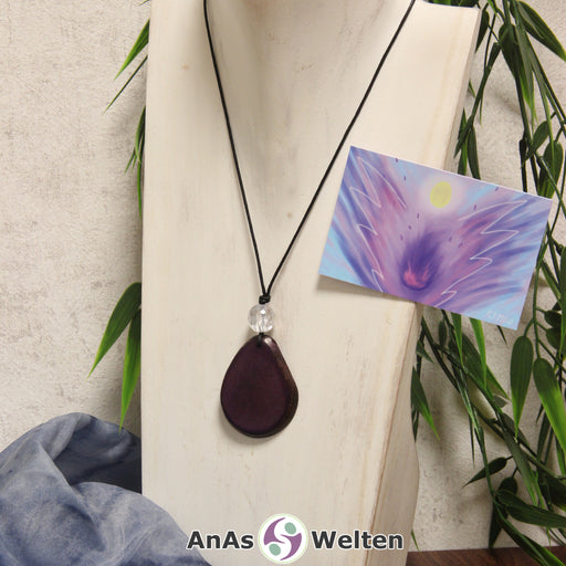 Das Produktbild für die Engel Kette Erzengel Zadkiel mit Karte zeigt eine in einem dunklen Lila gefärbte Tagua-Nuss-Scheibe an einem schwarzen Baumwollband. Die Nuss-Scheibe hat einen dunklen Rand und eine glänzende Oberfläche. Direkt darüber befindet sich eine facettierte Bergkristallkugel. Rechts neben der Kette sieht man außerdem die Erzengel Zadkiel Karte, die den Engel als abstraktes Bild zeigt.