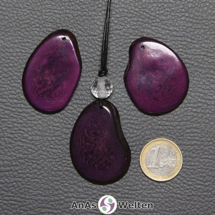 Drei Tagua-Nuss-Scheiben in Lila. Die mittlere ist mit einer Bergkristallkugel an ein schwarzes Baumwollband gebunden.