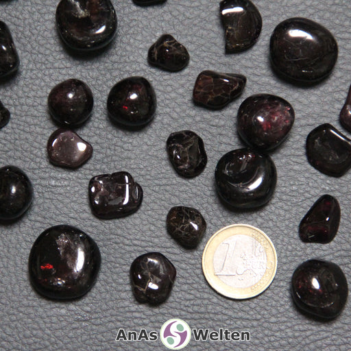 Das Bild zeigt den Granat Trommelstein als mehrere Beispielsteine. Die Edelsteine sind dunkelrot bis schwarz und haben gelegentliche leuchtend rote Stelle. Sie sind zwar geschliffen, haben aber trotzdem gelegentlich eine unebene Form.