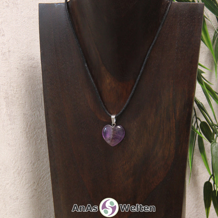 Ein Amethyst Herz Anhänger an einem schwarzen Baumwollband, der um eine dunkle Büste gehängt wurde.
