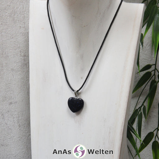 Das Bild zeigt einen Herzanhänger Blaufluss-Herz an einem schwarzen Baumwollband. Der Edelstein wurde in eine Herzform geschliffen und verfügt über eine silberfarbene Öse an der Oberseite. Er hat eine tiefblaue Farbe, die von einem silbrigen und blauen Glitzern durchzogen ist.