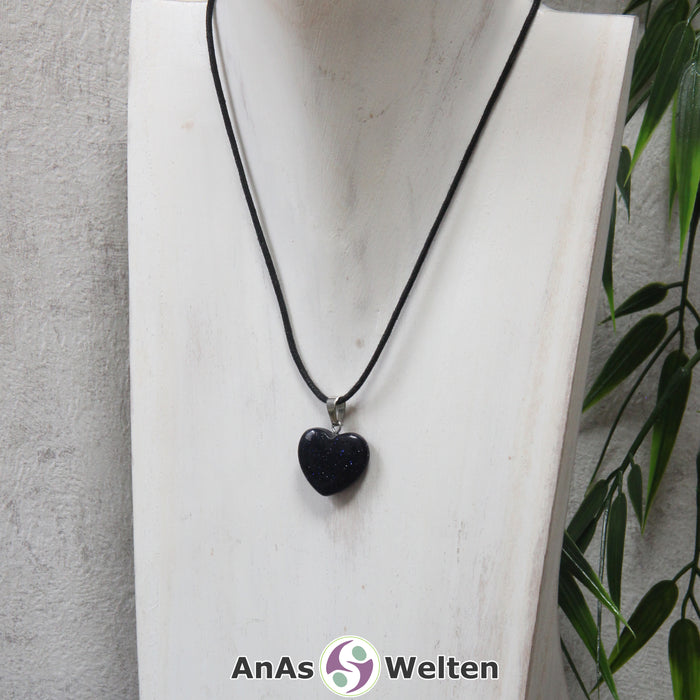Das Bild zeigt einen Herzanhänger Blaufluss-Herz an einem schwarzen Baumwollband. Der Edelstein wurde in eine Herzform geschliffen und verfügt über eine silberfarbene Öse an der Oberseite. Er hat eine tiefblaue Farbe, die von einem silbrigen und blauen Glitzern durchzogen ist.