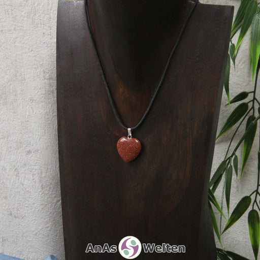 Das Bild zeigt einen Herzanhänger Goldfluss-Herz an einem schwarzen Baumwollband. Der Edelstein wurde in eine Herzform geschliffen und verfügt über eine silberfarbene Öse an der Oberseite. Er hat eine rotbraune Farbe, die von einem goldenen Glitzern durchzogen ist.