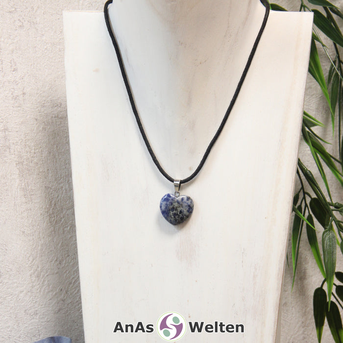 Das Bild zeigt einen Herzanhänger Sodalith-Herz an einem schwarzen Baumwollband. Der Edelstein wurde in eine Herzform geschliffen und verfügt über eine silberfarbene Öse an der Oberseite. Er hat ein unregelmäßiges Muster in den Farben Blau und Weiß.