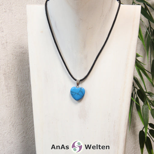 Das Bild zeigt einen Herzanhänger Turquenit-Herz an einem schwarzen Baumwollband. Der Edelstein wurde in eine Herzform geschliffen und verfügt über eine silberfarbene Öse an der Oberseite. Er hat gleichmäßige türkise Farbe, die von dunkelgrauen Linien durchzogen ist.