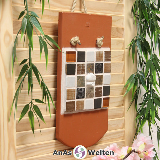 Das Bild zeigt ein Sator Quadrat Mosaik auf einem Biberschwanzdachziegel. Es ist mit einem Seil an einem Haken an der Wand aufgehängt. Die Fliesen sind braun, grau, weiß, golden und silbergrau. Dort, wo im Sator Quadrat die Buchstaben stehen, sind viereckige Fliesen zu sehen, die je eine Farbe oder ein anderes Muster pro unterschiedlichen Buchstaben haben. In der Mitte, wo normalerweise das N steht, sind zwei Magnesite in den Fugenmörtel eingesetzt.