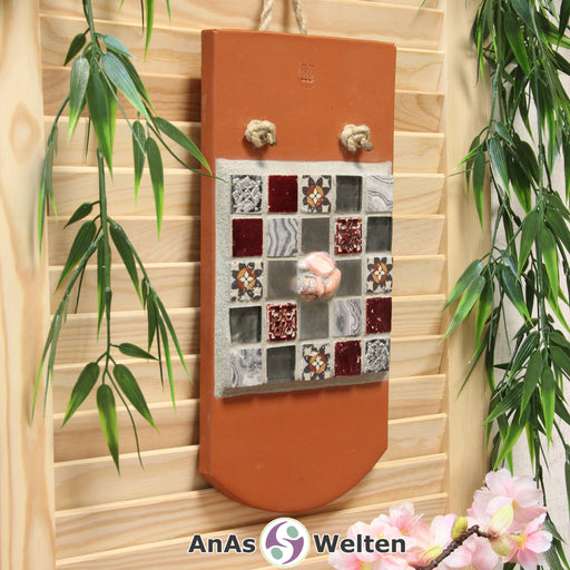 Das Bild zeigt ein Sator Quadrat Mosaik auf einem Biberschwanzdachziegel. Es ist mit einem Seil an einem Haken an der Wand aufgehängt. Die Fliesen sind silberfarben, rot, rot-grau in Vintageoptik. Dort, wo im Sator Quadrat die Buchstaben stehen, sind viereckige Fliesen zu sehen, die je eine Farbe oder ein anderes Muster pro unterschiedlichen Buchstaben haben. In der Mitte, wo normalerweise das N steht, sind drei rosa Achate in den Fugenmörtel eingesetzt.