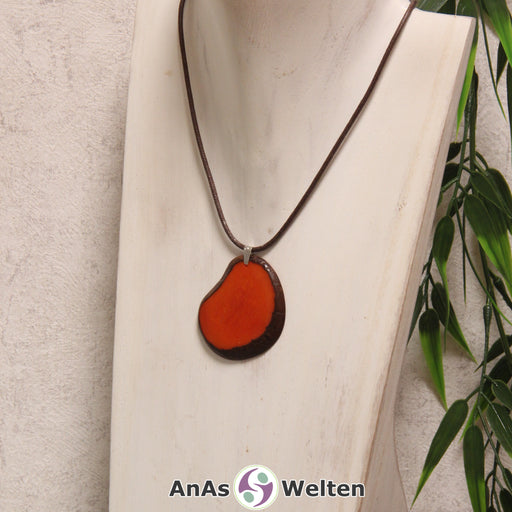 Das Produktbild zeigt die Tagua-Nuss-Schmuck Kette orange. Zu sehen ist eine orangene Tagua-Nuss-Scheibe, die mit einer Edelstahlöse an einem braunen Baumwollband befestigt ist. Die Nuss-Scheibe hat einen dunklen Rand und eine glänzende Oberfläche.