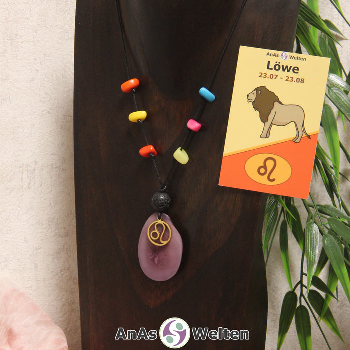 Eine lila Tagua-Nuss Sternzeichen Kette mit bunten Taguasplittern, goldenem Löwe-Symbol und Lavakugel an einer dunklen Büste.