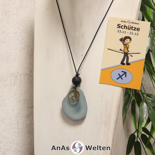 Das Produktbild für die Tagua-Nuss-Schmuck Sternzeichen-Kette Schütze zeigt eine in himmelblau gefärbte Tagua-Nuss-Scheibe an einem schwarzen Baumwollband, vor der ein goldfarbener Anhänger mit dem Sternzeichensymbol des Schützen befestigt ist. Die Nuss-Scheibe hat einen dunklen Rand und eine glänzende Oberfläche. Direkt darüber befindet sich eine Lavakugel. Rechts neben der Kette sieht man außerdem die Sternzeichen Schütze Karte von AnAs Welten.