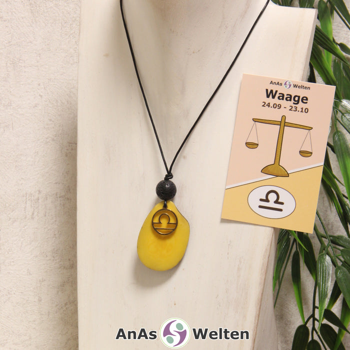 Eine gelb gefärbte Tagua-Nuss Sternzeichen Kette mit goldfarbenem Waage-Symbol und Lavakugel an einer hellen Büste.