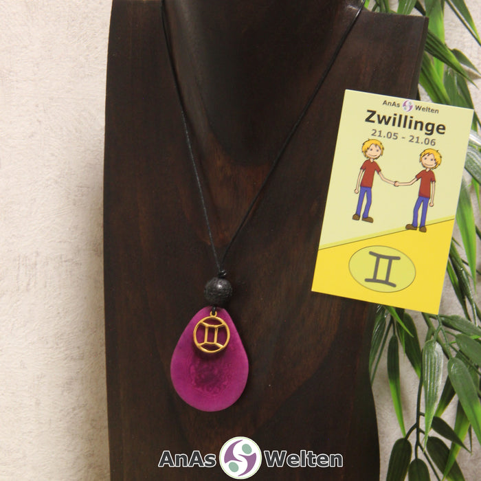 Eine violett gefärbte Tagua-Nuss Sternzeichen Kette mit goldfarbenem Zwillinge-Symbol und Lavakugel an einer dunklen Büste.