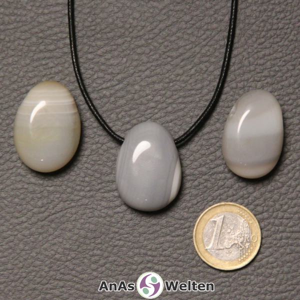 Drei grau-weiße Achat gebohrte Trommelsteine nebeneinander. Einer befindet sich an einem schwarzen gewachsten Baumwollband.