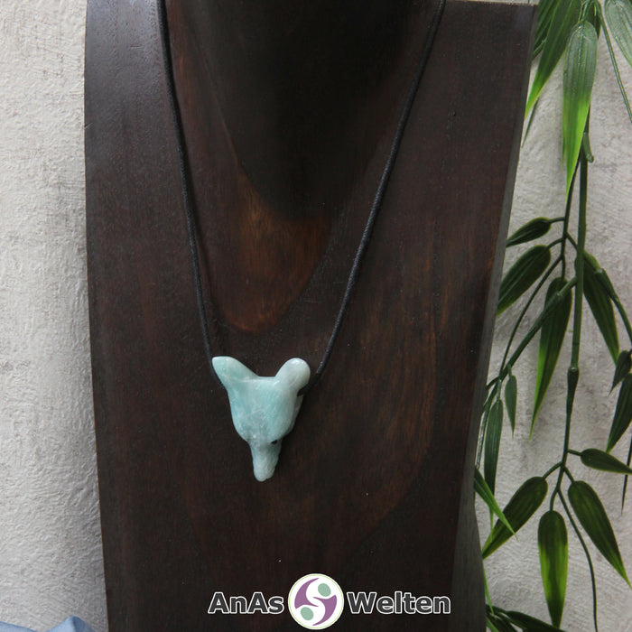 Das Bild zeigt einen Amazonit Wolf Anhänger an einem Baumwollband. Der Edelstein hat eine hellblaue Farbe mit einer türkis-blauen Musterung. Außerdem sind in seinem Inneren schwach weiße Risse zu erkennen. Das Fellmuster und die Nase sind durch kleine Kerben im Edelstein dargestellt. Die Augen sind kleine schwarze Perlen.