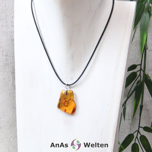 Das Bild zeigt einen Bernstein Anhänger Sternzeichen Stier mit Silberöse an einem Baumwollband. Der Bernstein ist flach, an den ungeschliffenen Kanten poliert und hat die typische honiggoldene Farbe von Bernstein. Da er durchsichtig ist, kann man deutlich das dunkel auf die Rückseite eingravierte Sternzeichensymbol des Stiers erkennen.