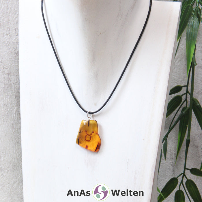 Das Bild zeigt einen Bernstein Anhänger Sternzeichen Stier mit Silberöse an einem Baumwollband. Der Bernstein ist flach, an den ungeschliffenen Kanten poliert und hat die typische honiggoldene Farbe von Bernstein. Da er durchsichtig ist, kann man deutlich das dunkel auf die Rückseite eingravierte Sternzeichensymbol des Stiers erkennen.