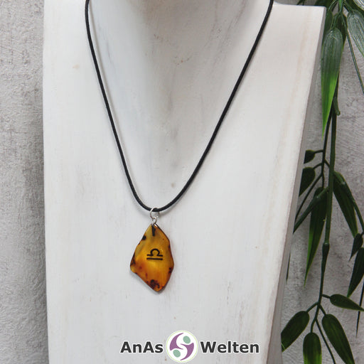 Das Bild zeigt einen Bernstein Anhänger Sternzeichen Waage mit Silberöse an einem Baumwollband. Der Bernstein ist flach, an den ungeschliffenen Kanten poliert und hat die typische honiggoldene Farbe von Bernstein. Da er durchsichtig ist, kann man deutlich das dunkel auf die Rückseite eingravierte Sternzeichensymbol der Waage erkennen.