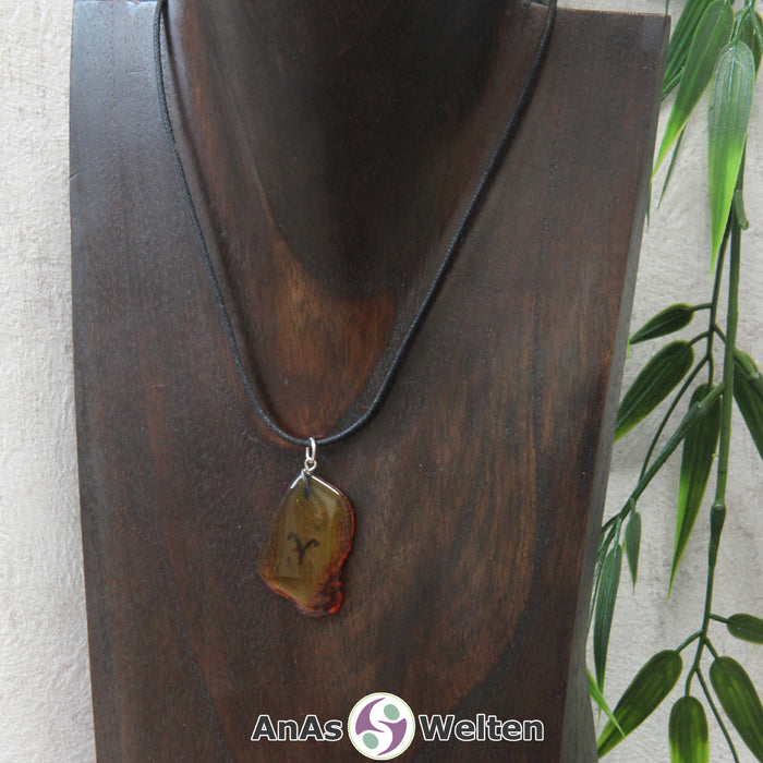 Ein Bernstein Anhänger Widder an einem Baumwollband als Kette um eine dunkle Büste.