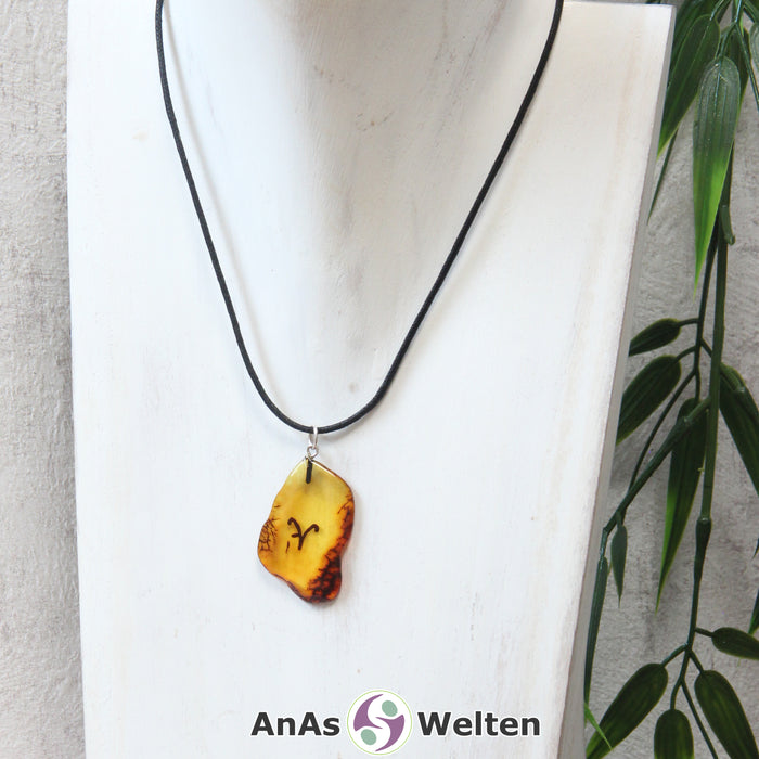 Das Bild zeigt einen Bernstein Anhänger Sternzeichen Widder mit Silberöse an einem Baumwollband. Der Bernstein ist flach, an den ungeschliffenen Kanten poliert und hat die typische honiggoldene Farbe von Bernstein. Da er durchsichtig ist, kann man deutlich das dunkel auf die Rückseite eingravierte Sternzeichensymbol des Widders erkennen.