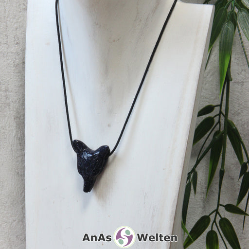 Das Bild zeigt einen Blaufluss Wolf Anhänger an einem Baumwollband. Der Edelstein hat ein tiefes Dunkelblau, das von unzähligen golden glitzernden Punkten durchzogen ist. Das Fellmuster und die Nase sind durch kleine Kerben im Edelstein dargestellt. Die Augen sind kleine schwarze Perlen.