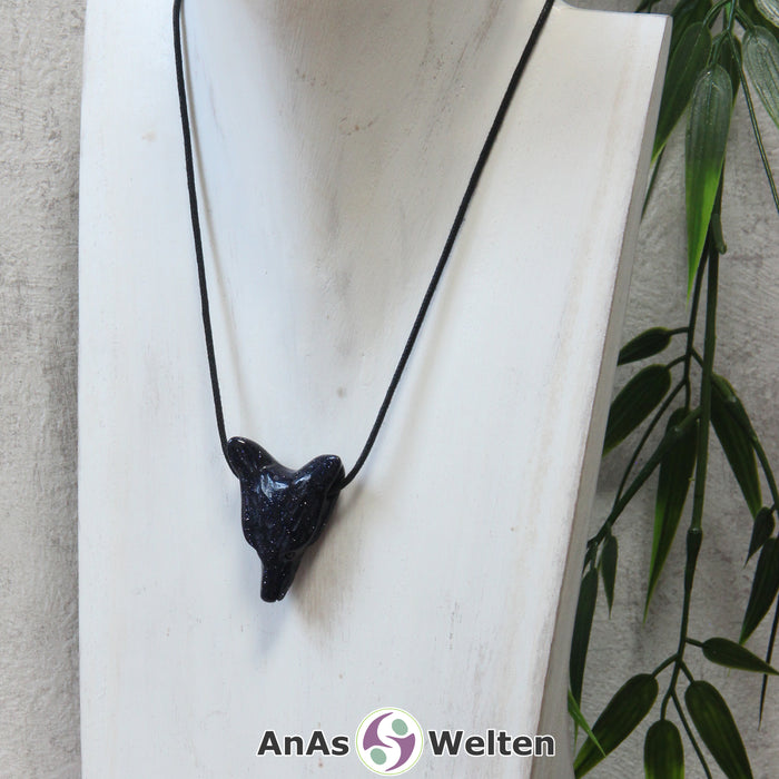 Das Bild zeigt einen Blaufluss Wolf Anhänger an einem Baumwollband. Der Edelstein hat ein tiefes Dunkelblau, das von unzähligen golden glitzernden Punkten durchzogen ist. Das Fellmuster und die Nase sind durch kleine Kerben im Edelstein dargestellt. Die Augen sind kleine schwarze Perlen.