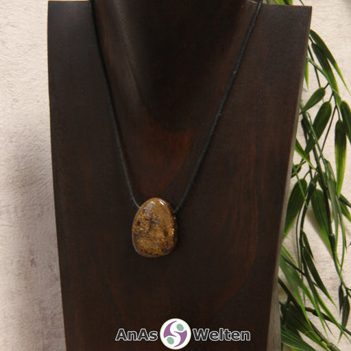 Ein gebohrter Bronzit Trommelstein an einem Baumwollband. Der Edelstein hat eine bronzeähnliche Färbung und ein unregelmäßiges Muster aus Brauntönen.